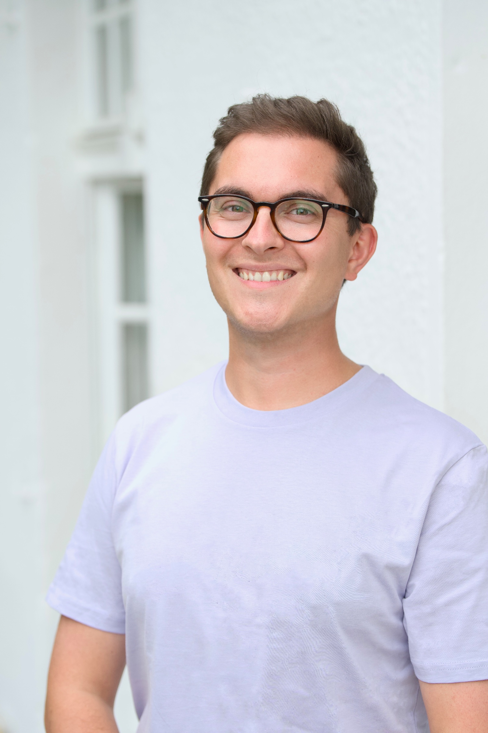 Gabi, a young man with short brown hair and dark round glasses, smiling warmly at the camera. He wears a light lavender t-shirt and stands in front of a white painted building.